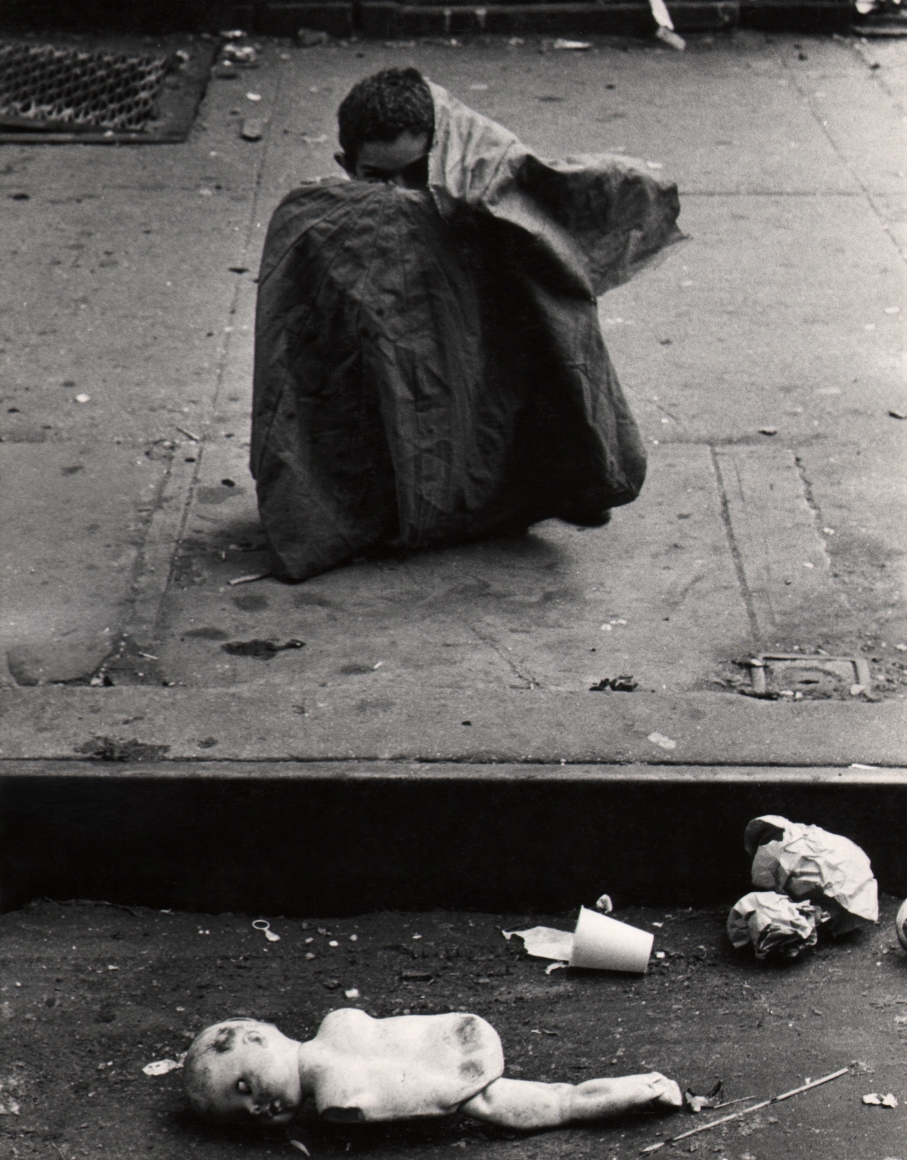 34. Beuford Smith, Boy & Doll, Lower East Side, NYC, ​1966. A young boy sits on the sidewalk with most of his body covered by some sort of cloth; a broken doll and other trash is on the street in front of him at the bottom of the frame.