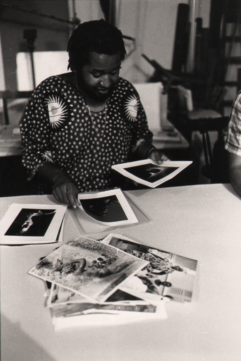 01. Anthony Barboza, Shawn Walker, International Black Photographers Annual, 1973. A man in a patterned shirt flips through a stack of photographs on a white table.