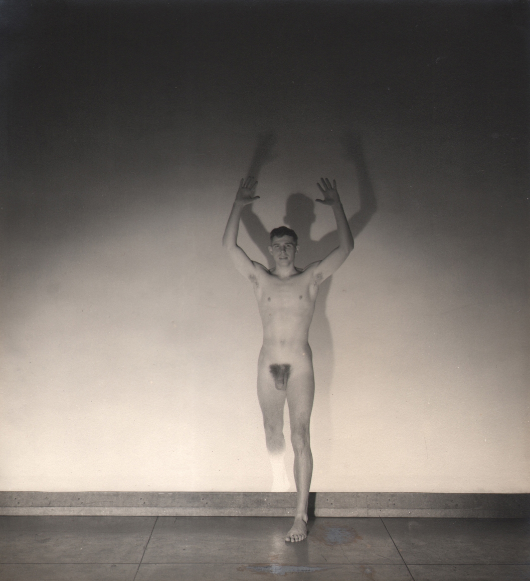 George Platt Lynes, Jeremy Jackson, ​1935. Male nude figure stands with arms and one leg raised against a spot-lit wall.