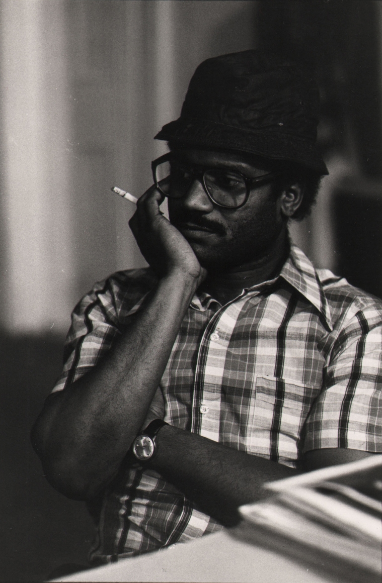 03. Anthony Barboza, Beuford Smith, First Black Photographers Annual Meeting, NYC, ​1970s. Torso of a seated man in a checked shirt wearing glasses and a hat, one hand held to his face.
