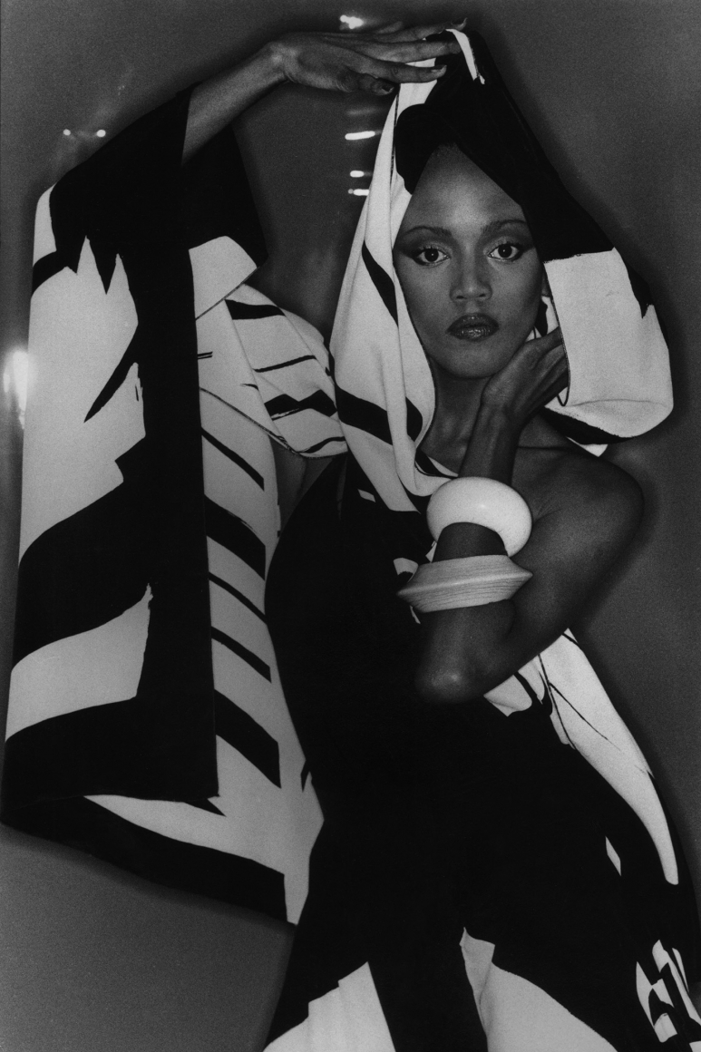 04. Anthony Barboza, Toukie Smith, ​1980s. Model stands in a draping, graphic, hooded garment with one arm above her head and one raised to her face.