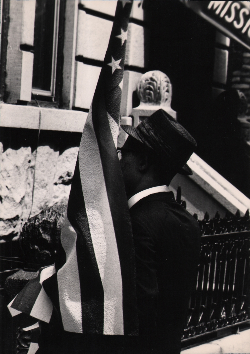 22. Shawn Walker, Untitled, ​c. 1965. Man in a jacket and hat carries an American flag, facing away from the photographer.