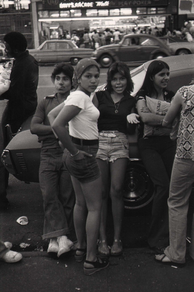 11. Anthony Barboza, Coney Island, NY, 1970s. A group of young people socializing by a parked car. The central figure looks to the camera in a white t-shirt with hands in her back shorts pockets.