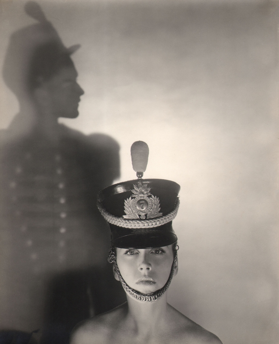 George Platt Lynes, Bridget Chisholm, ​c. 1945. Model in a hat pictured from shoulders up looking into the camera. A silhouetted figure in profile to the left in the background.