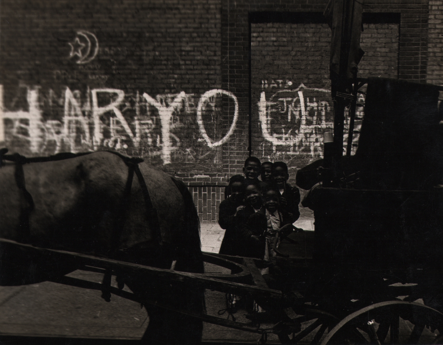 23. Shawn Walker, HarYourAct, ​1963. A group of six children stand behind a horse-drawn carriage in the foreground. The background is a graffiti-covered brick wall.