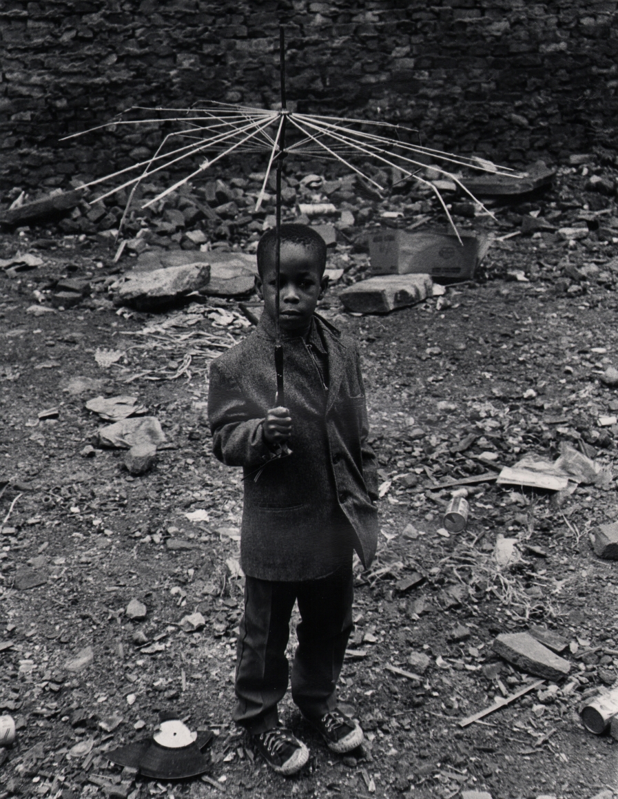 36. Beuford Smith, Boy with Umbrella, ​1973. A young boy holds a broken umbrella, with only wires remaining, above his head.