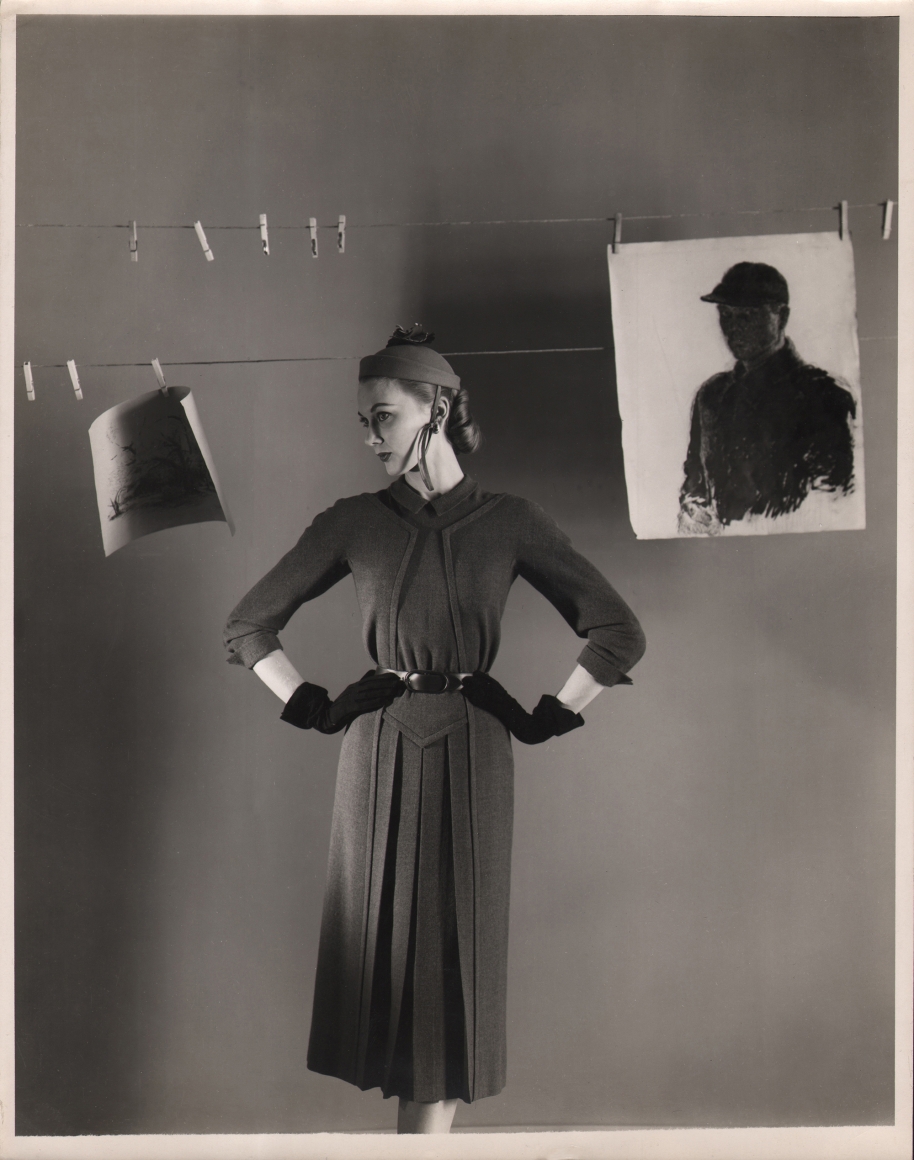 George Platt Lynes, Henri Bendel, c. 1950. Model stands with hands on hips, head turned to the left, beneath a clothes line with two drawings suspended from it.
