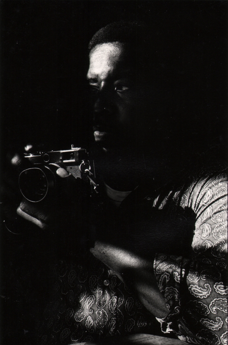 08. Anthony Barboza, Lou Draper, ​1970s. Dimly lit torso of a man facing left and holding a camera at chest-level.