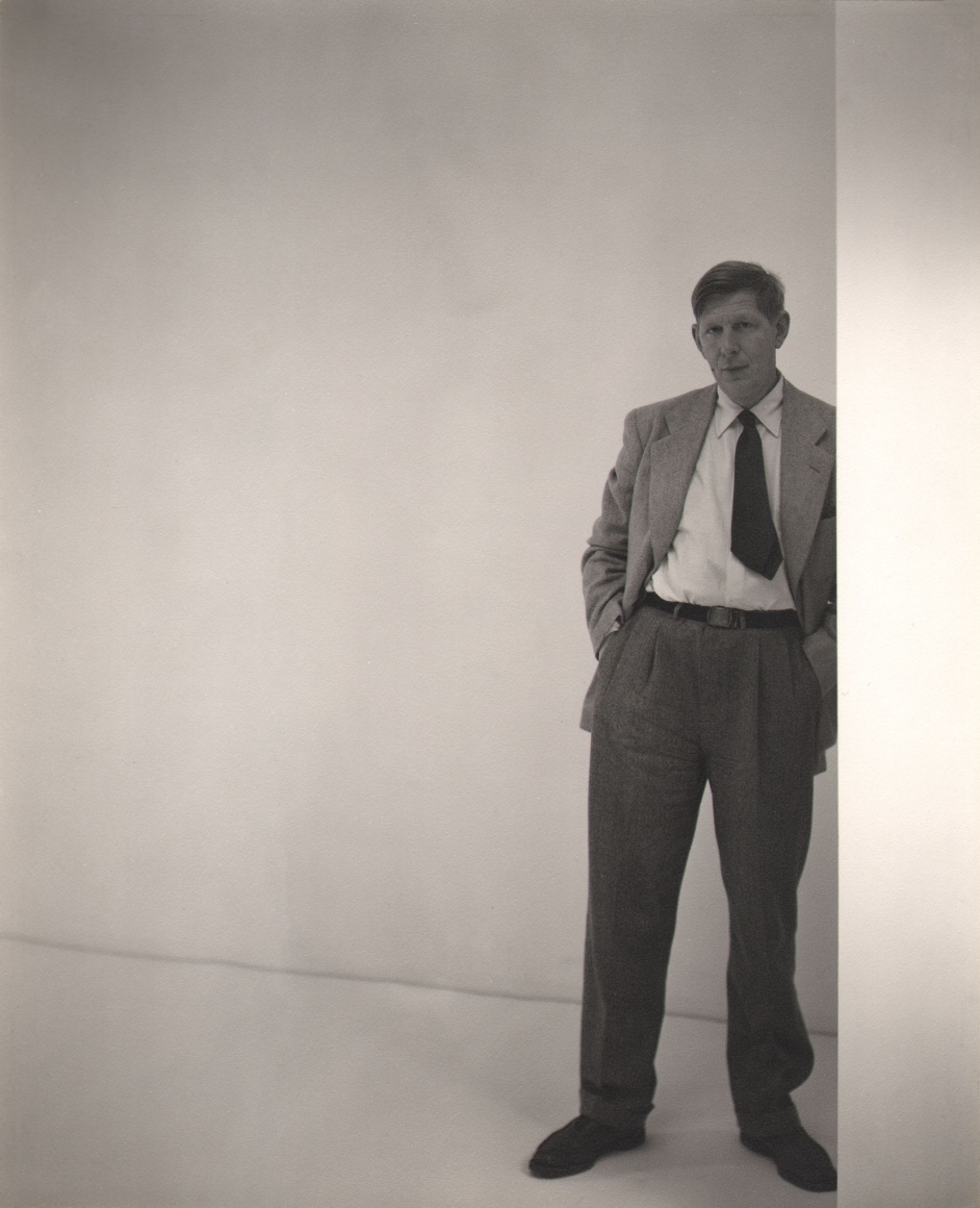 George Platt Lynes, W.H. Auden, ​1947. Subject stands on the right of the frame leaning against a white wall.