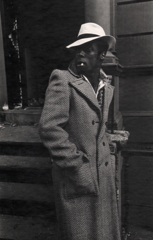 09. Anthony Barboza, Harlem, NY, ​1970. A man standing on the sidewalk with hands in a long coat. He wears a white hat, body facing the right with his face turned left.