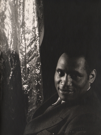 Carl Van Vechten, Paul Robeson, ​1933. Subject smiles to the camera with torso facing left, a reflective material hangs to the left of the frame.