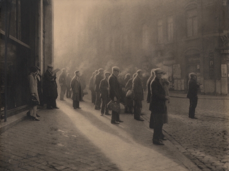 08. Léonard Misonne, Première cigarette, 1927. Various figures, mostly capped men, stand on the sidewalk, cigarette smoke rising above them. Sepia-toned print.