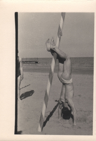 PaJaMa, Untitled, c. 1945. A man in a swimsuit on the beach does a handstand with legs against a striped pole.