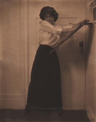 01. Gertrude Käsebier, Untitled, ​c. 1910. A woman in a white shirt and long black skirt standing in front of a door, seeming to draw against a wooden easel.