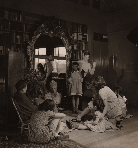 Louise Dahl-Wolfe, Charades Before Bedtime, Editorial in Diana Vreeland's House, 1942. A group of children sits on the floor looking toward two standing children. One has hands raised in black gloves and facepaint, the other stands behind her pointing a thumb to the right of the frame.
