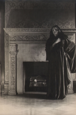 PaJaMa, Margaret French, Hoboken, ​c. 1945. A woman stands wearing draping fabric in front of a fireplace. One hand is on her hip and the other held to her chest.