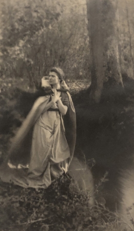 07. Dorothy & Reta Morter, Destiny - Self-Portrait, ​c. 1918. A woman stands in the left of the frame by a river in a dress and cloak, looking to the left.
