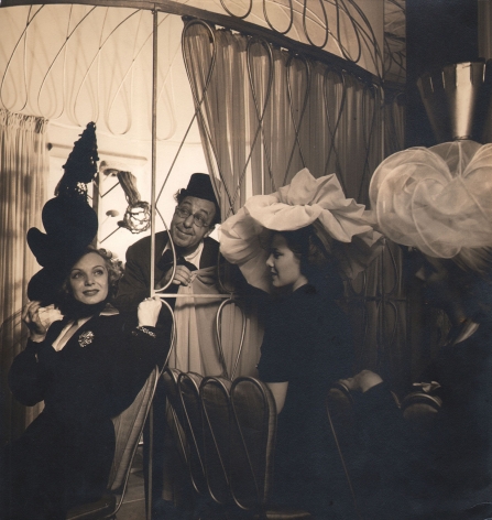 Louise Dahl-Wolfe, Ed Wynn & Jane Pickens In Boys & Girls Together, 1940. A man in glasses and a top hat peers out from behind a curtain. Three women in large billowing hats are seated in the foreground.