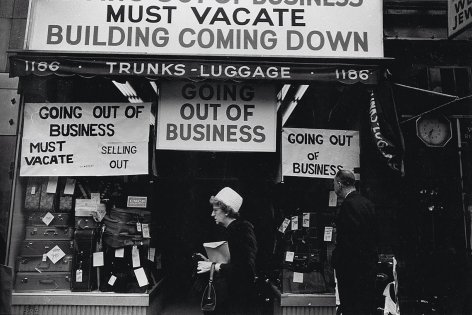 7. Simpson Kalisher, Untitled, ​1962. Two pedestrians pass a storefront with various "Going out of business" signs and stacks of suitcases in the shop window.
