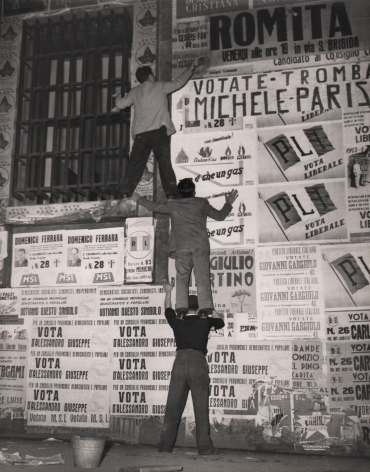 Federico Garolla, Elezione a Napoli, ​1948. Three men stand on each others shoulders to reach a high point on a wall covered in election posters.