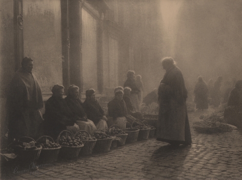 07. Léonard Misonne, Choix difficile, 1933. Older woman stands on a cobbled street facing a row of women seated with baskets of produce. Sepia-toned print.