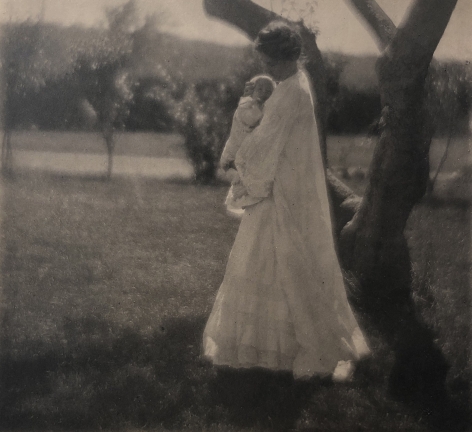 04. Gertrude Käsebier, Blossom Day, ​1904. A woman in a long white dress stands beneath a tree holding a baby to her chest.