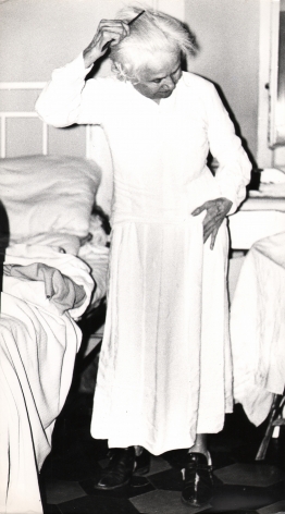 01. Mario Giacomelli, Verrà la morte e avrà i tuoi occhi, 1966–1968. High contrast image. An old woman stands by a bed, combing her hair.