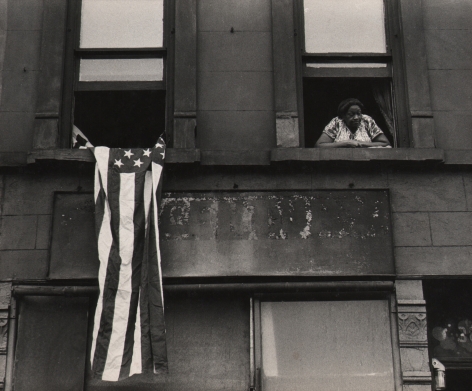 09. Beuford Smith, Flag Day, Harlem, ​1976. Detail of two windows of an apartment building; a woman leans out of the right window, an American flag hangs from the left.