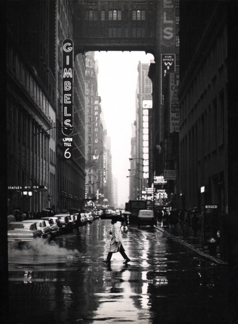 10. Jan Lukas, New York, 32nd Street, ​1964. A man with a trenchcoat and umbrella walks across a wet car-lined street. The neon sign for Gimbels is on the left midground.
