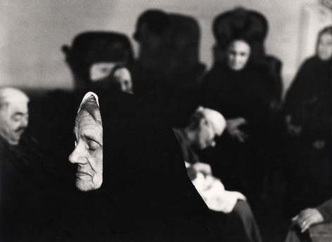06. Mario Giacomelli, Verrà la morte e avrà i tuoi occhi, 1966–1968. High contrast image. A cloaked woman in the foreground with closed eyes, facing the left of the frame. Various figures out of focus behind her.
