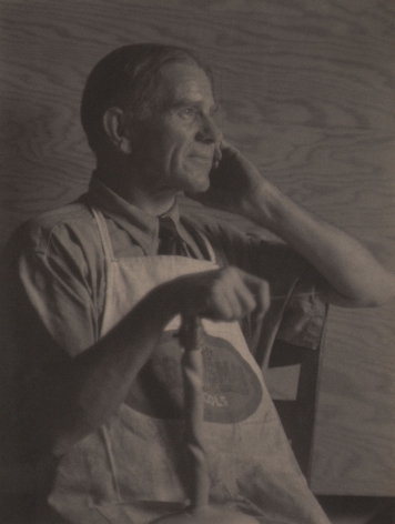 Doris Ulmann, Park Fisher, Brasstown, N.C. (Wood carver), 1928–1934. Seated man in an apron smiling off to the right of the frame with one hand held to his face and the other resting on a carved wooden object in front of him.