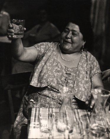 Carlo Bavagnoli, Gente di Trastevere, ​1957–1958. A smiling woman seated at a table raises her drinking glass in a toast.