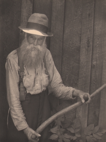 10. Doris Ulmann, Untitled (Farmer holding oxen yoke), 1928–1934. Older bearded man in a hat standing against a wooden wall and suspenders holding a wooden rod with two handles.