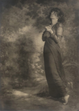 06. Mesdames Morter, Love Praying - Self-Portrait, ​1923. A woman stands in a dimly lit room in a long draping, dress with one hand on her hip, looking down.