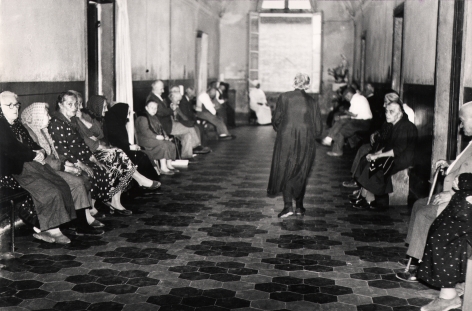 05. Mario Giacomelli, Verrà la morte e avrà i tuoi occhi, 1966–1968. High contrast image. A woman walks down a hallway, back to the photographer. The hall is lined with seated women.