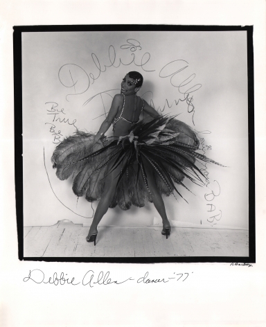 Anthony Barboza, Debbie Allen - Dancer, ​1977. Subject stands in feathered skirt, back to camera, legs wide. Head is turned smiling to camera. Writing on white backdrop includes her name.