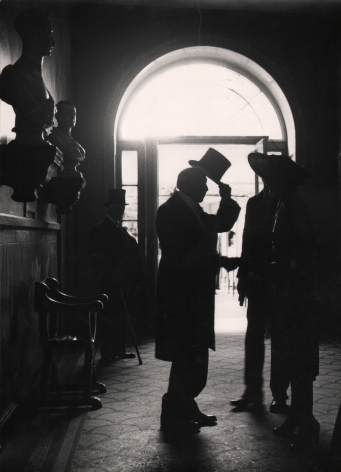 13. Lord Snowdon, The late Mr. William Stone, in Albany, London, c. 1966. Figures silhouetted in front of a doorway. Figure on the left lifts a tophat to two individuals facing him.