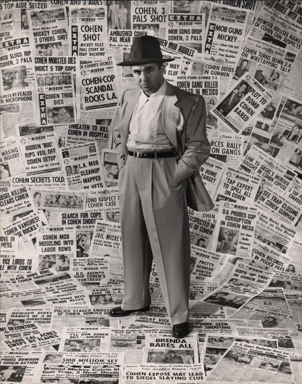 Ed Clark, Mickey Cohen, Gangster, 1949. A suited man stands against a backdrop of newspapers pasted to the wall and ceiling, hands in pockets, looking to the camera with a serious expression.