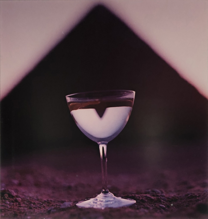 Bert Stern, Martini & Pyramid, for Smirnoff Vodka, ​1955. Martini glass on sand with a triangle shape out of focus in the background.