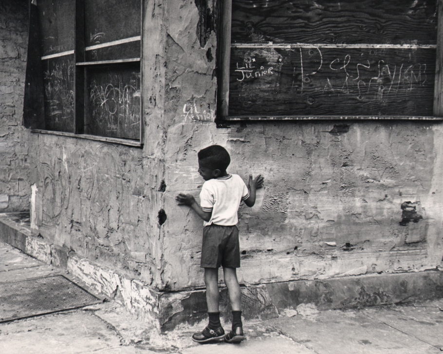 20. Beuford Smith, Playing 'Hide and Seek,' ​1968. A young boy stands against the corner of a run-down building.