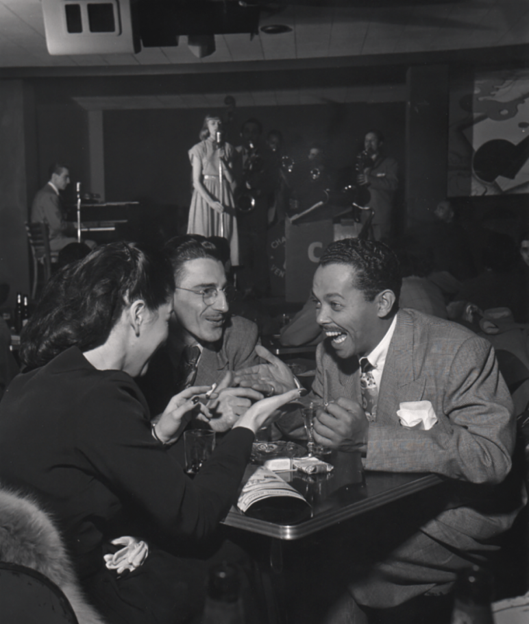 Wayne F. Miller, Billy Eckstine, ​1948. Subject is seated, smiling, at a club table with two other figures. A woman sings on stage in the background.